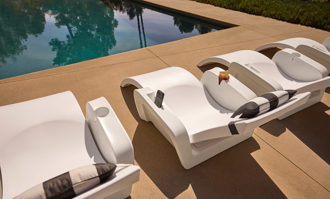 Three white poolside loungers with black-and-white pillows are lined up beside a clear swimming pool. One lounger holds a drink, and another has a smartphone in its holder.