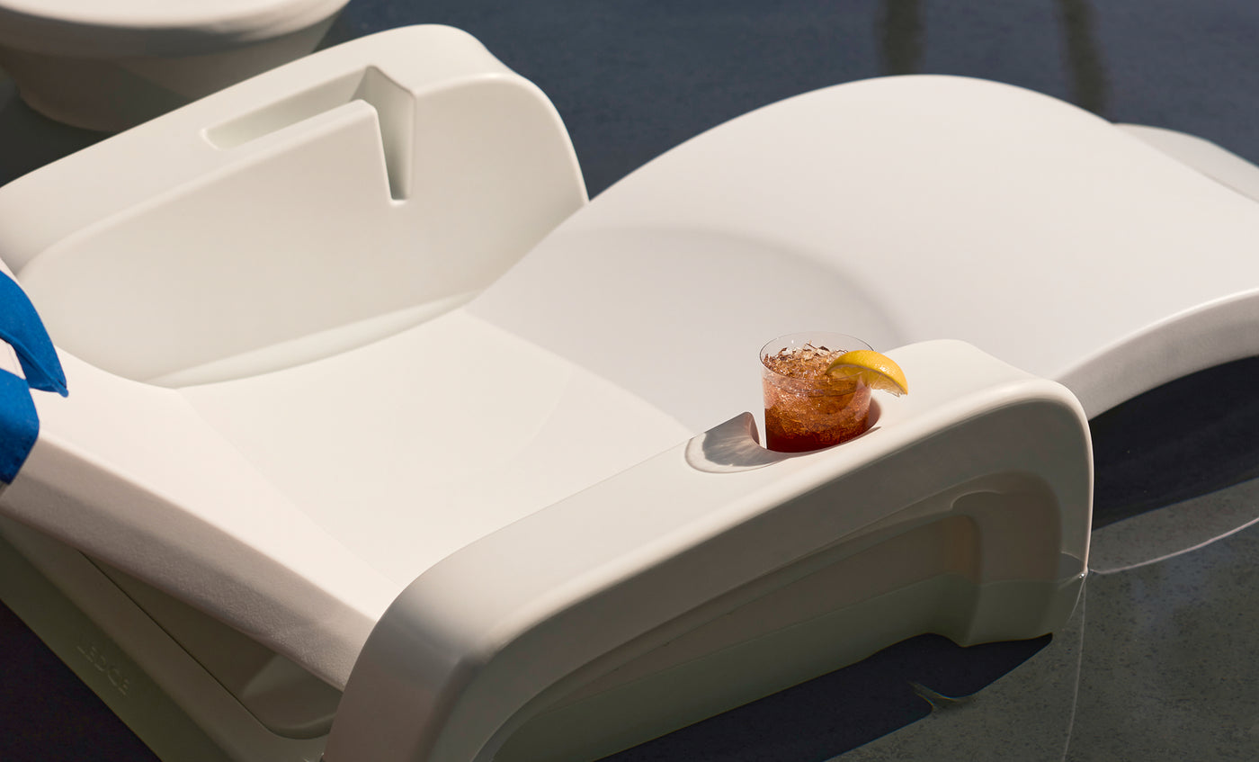 A white poolside lounge chair sits partially in water, holding a glass of iced drink with a lemon wedge on its armrest. Sunlight casts shadows, creating a relaxing poolside atmosphere.