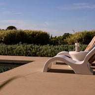 The Ledge Lounger Signature Lowback Chair in white sits poolside under a clear blue sky, surrounded by lush green bushes and trees on a sunny day, with a refreshing drink close by.