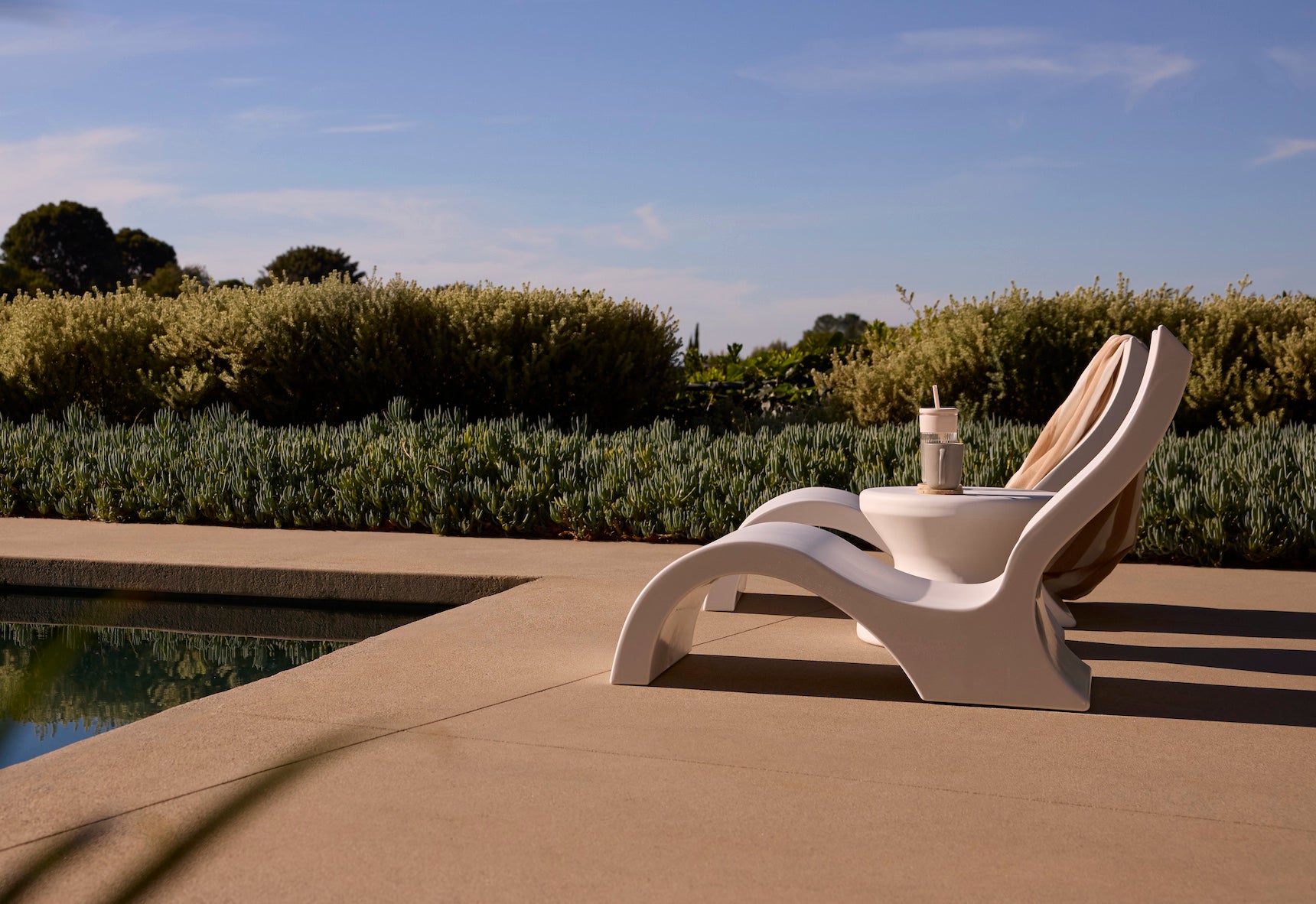 The Ledge Lounger Signature Lowback Chair in white sits poolside under a clear blue sky, surrounded by lush green bushes and trees on a sunny day, with a refreshing drink close by.