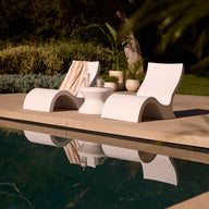Two Signature Lowback Chairs by Ledge Lounger and a small table with drinks and a towel sit poolside amid lush greenery, their reflections shimmering in the calm water—ideal outdoor seating for relaxing by the pool.