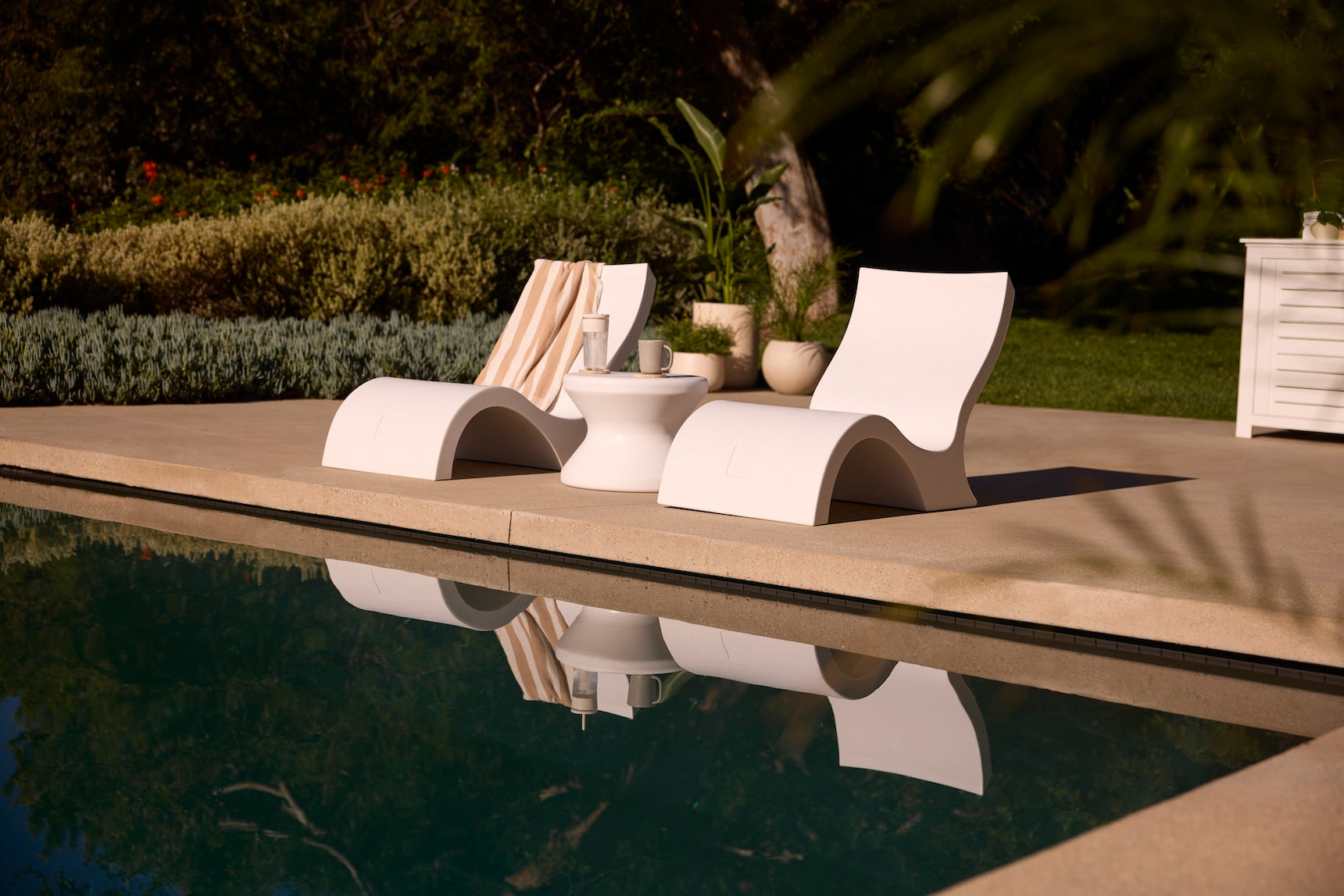 Two Signature Lowback Chairs by Ledge Lounger and a small table with drinks and a towel sit poolside amid lush greenery, their reflections shimmering in the calm water—ideal outdoor seating for relaxing by the pool.