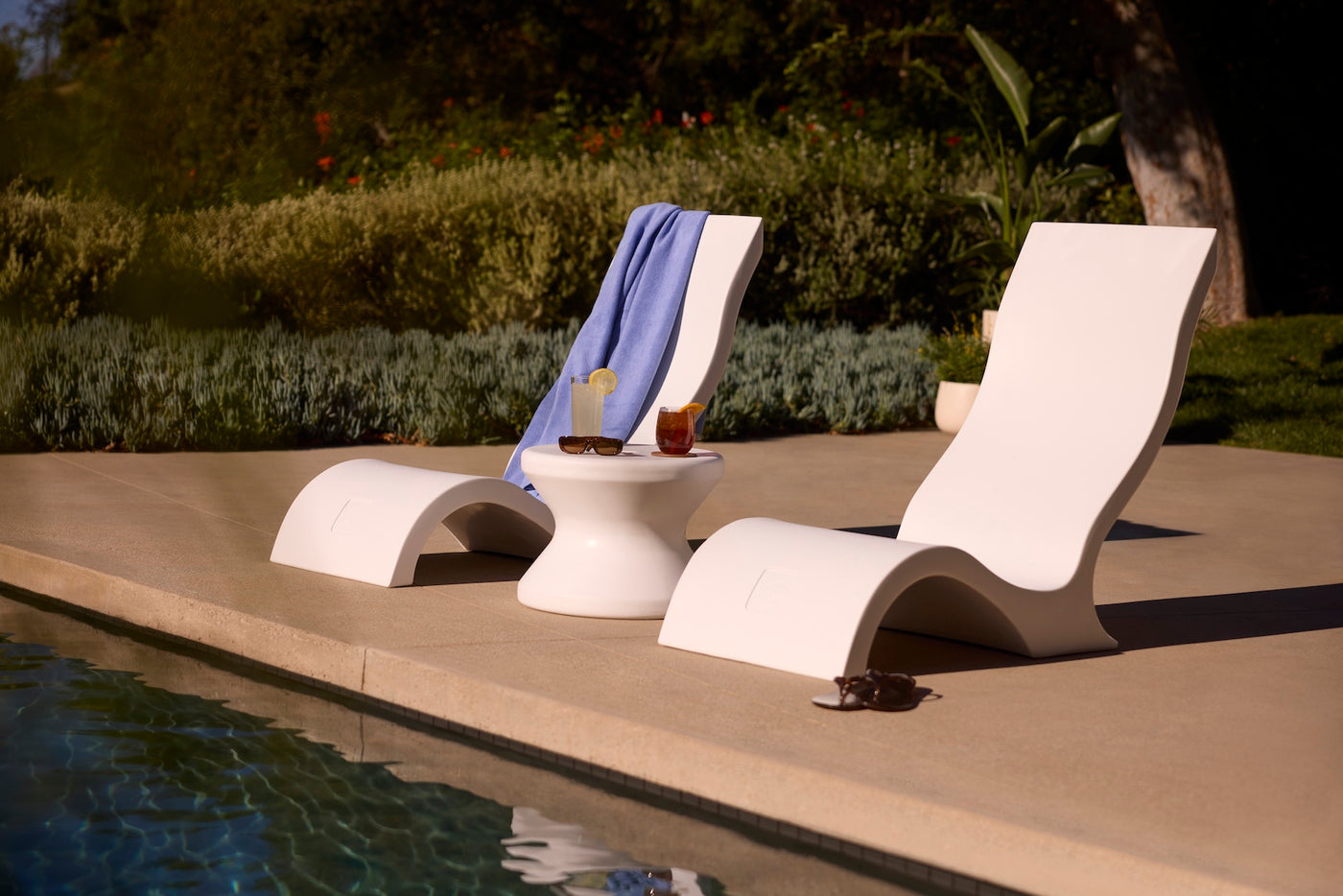 Two Signature Chairs by Ledge Lounger sit poolside with a small white table between them holding drinks. A blue towel is draped over one chair, while sunglasses rest nearby. Lush plants provide a vibrant, inviting backdrop.