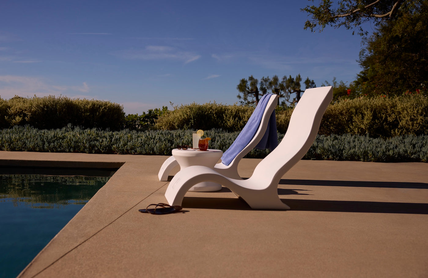 Two Ledge Lounger Signature Chairs with towels are set by a sparkling pool. A small table holds garnished drinks and sunglasses, surrounded by lush greenery under a blue sky for the ideal poolside setting.
