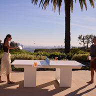 Two people enjoy a game with the Ledge Lounger Table Tennis and Barstools Gathering Set outside, drinks on the table, surrounded by greenery and palm trees, overlooking a cityscape under a clear sky.