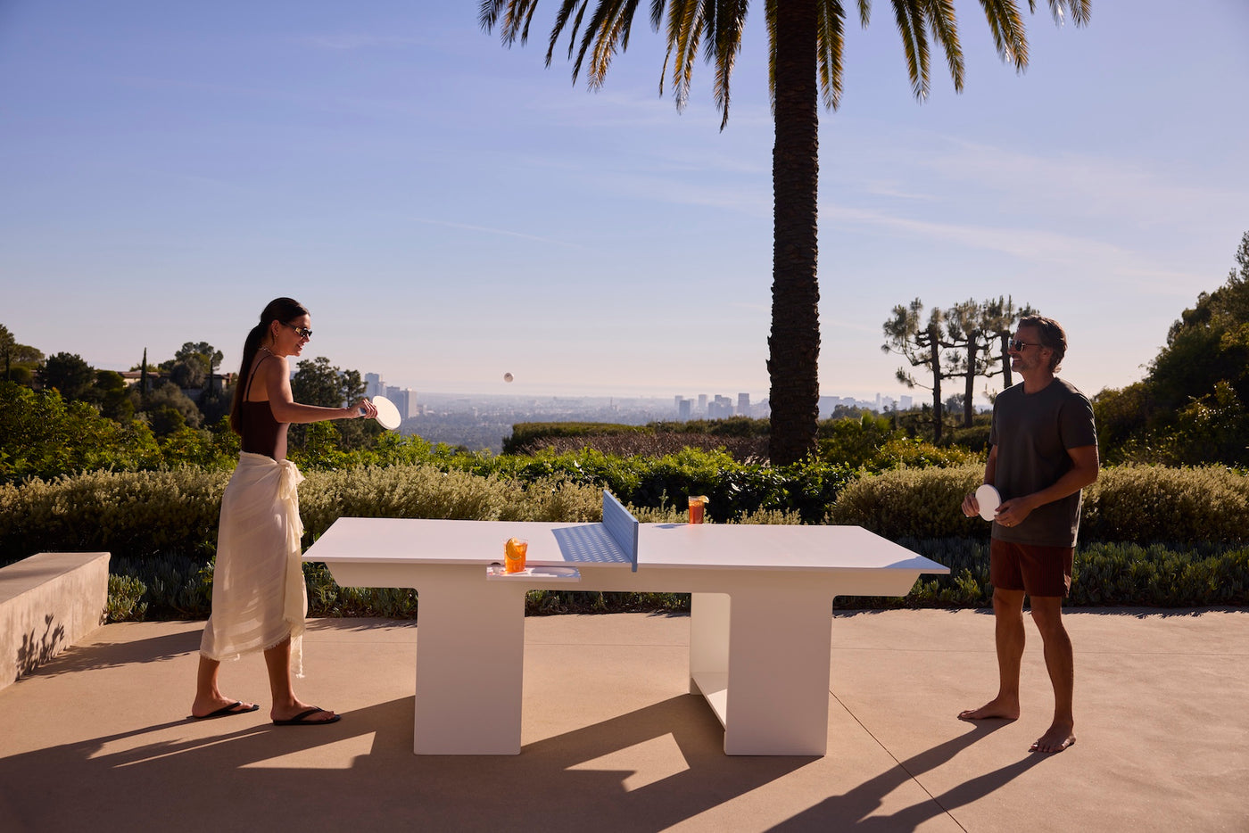 Two people enjoy a game with the Ledge Lounger Table Tennis and Barstools Gathering Set outside, drinks on the table, surrounded by greenery and palm trees, overlooking a cityscape under a clear sky.