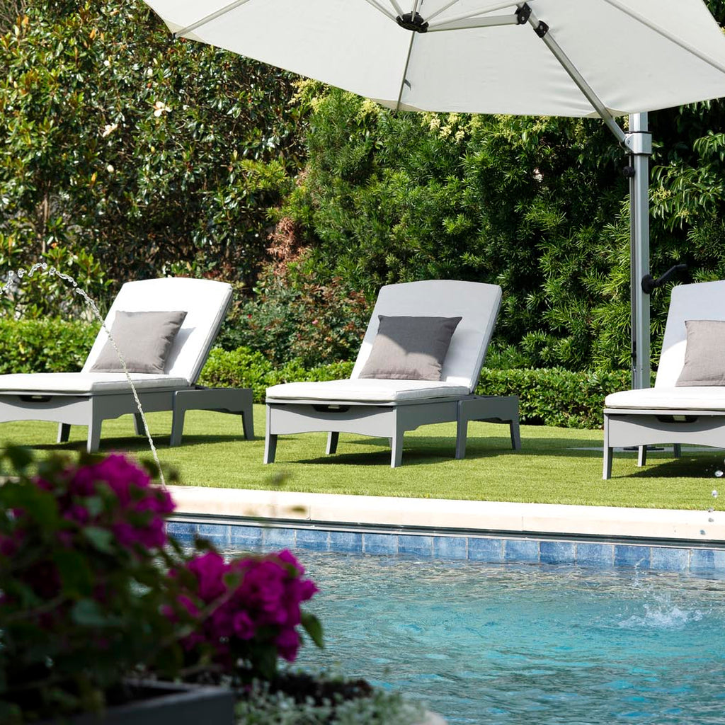 Three gray outdoor chaise lounges with white cushions under a large umbrella by a pool.
