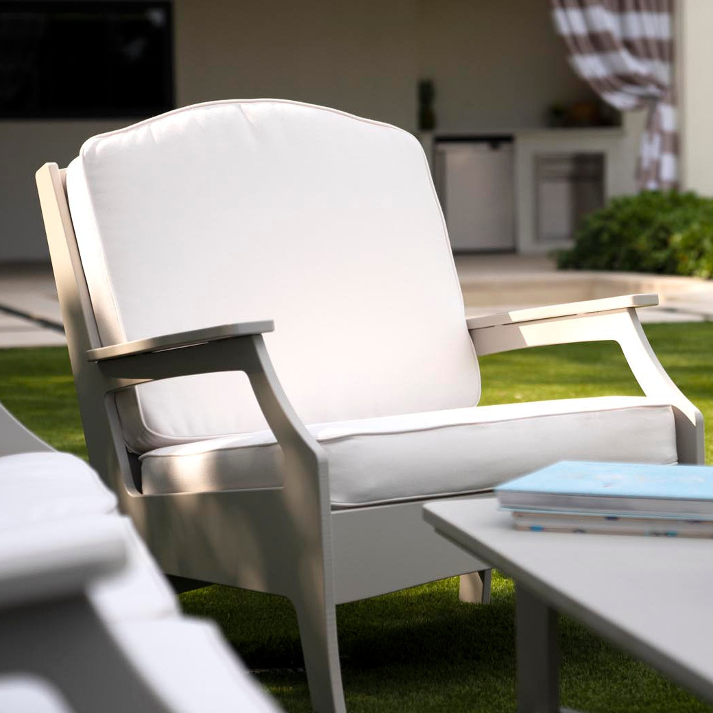 A white Legacy Club Chair from Ledge Lounger sits cushioned on the green lawn, creating a cozy outdoor space. Behind it, an outdoor kitchen with striped curtains adds charm, while a nearby table holds a book and a jug of iced tea, inviting relaxation and leisure in this customizable design.