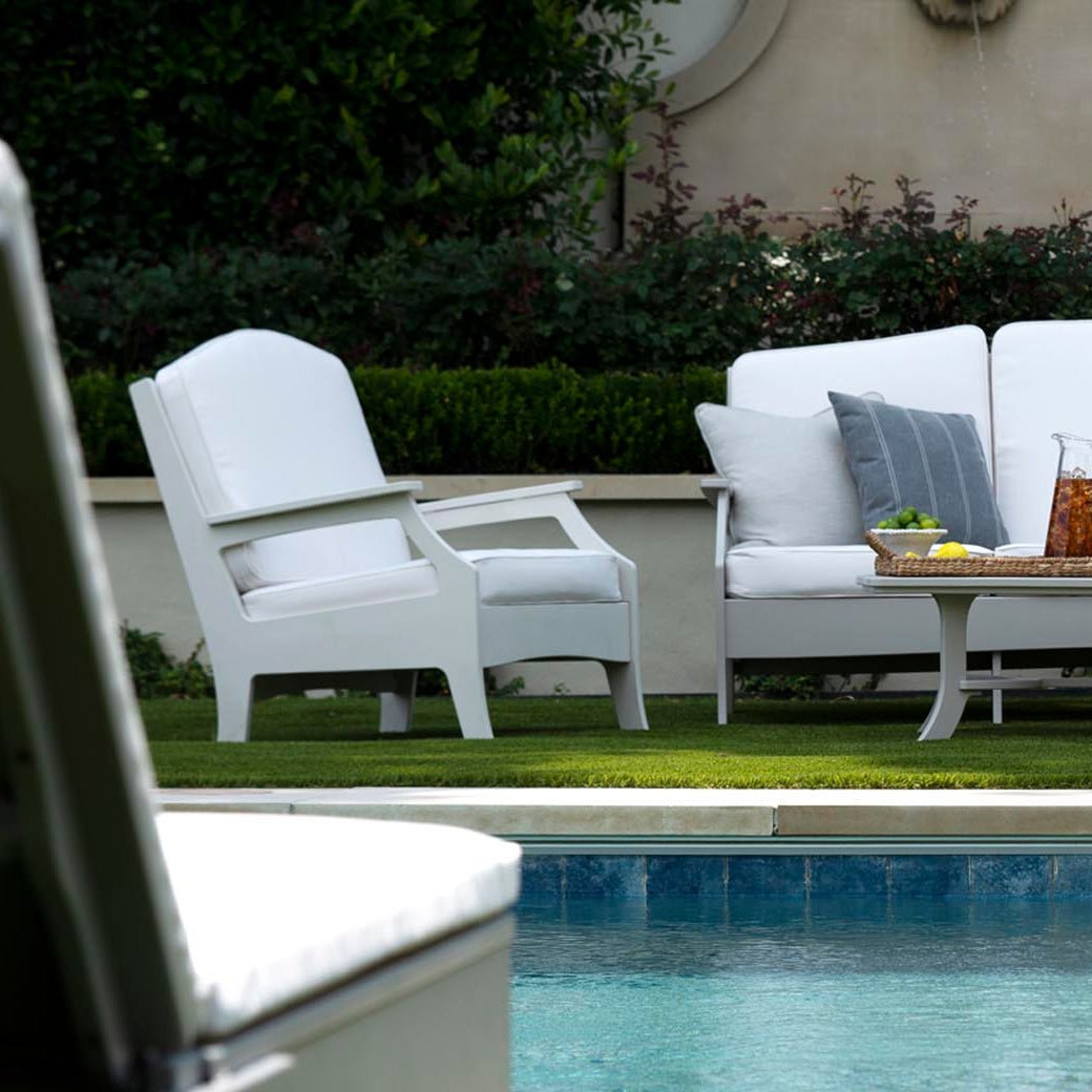 The serene poolside patio is furnished with elegant white lounge pieces, including a Legacy Club Chair by Ledge Lounger and a two-seater sofa. A table with iced tea adds to the scene, set against lush greenery and hedges for ultimate relaxation.