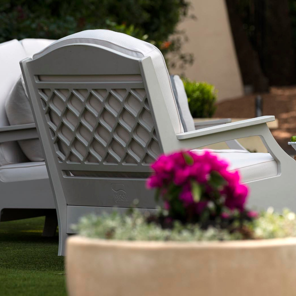 A cozy outdoor space boasts a pair of plush Legacy Club Chairs by Ledge Lounger, positioned around a low table with iced tea, glasses, grapes, and lemons. A large planter filled with vibrant pink flowers completes this inviting scene.