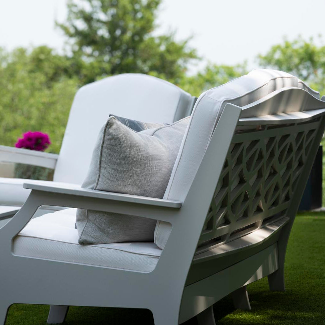 Legacy Club Chairs with cushions face a white coffee table on a grass lawn. A pitcher of iced tea and a snack bowl rest on the table. Potted purple flowers are nearby, framing the inviting outdoor space with blurred trees and a cityscape in the background. Brand: Ledge Lounger.