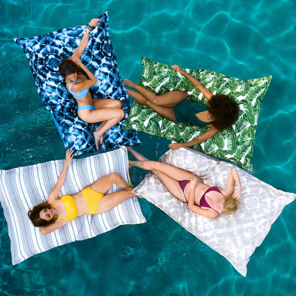 Four women are lounging on Laze Pillow floats by Ledge Lounger in a clear blue pool. Theyre wearing swimsuits with eyes closed, their colorful floats arranged in a cross shape, enjoying unparalleled outdoor comfort as they drift peacefully on the calm, vibrant water.