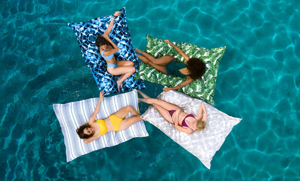 Four women are lounging on Laze Pillow floats by Ledge Lounger in a clear blue pool. Theyre wearing swimsuits with eyes closed, their colorful floats arranged in a cross shape, enjoying unparalleled outdoor comfort as they drift peacefully on the calm, vibrant water.