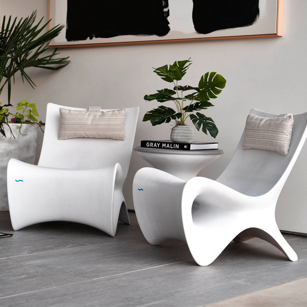A modern interior features two sleek white chairs with beige cushions, centered around a small white table holding a stack of books and a potted plant. Large potted greenery sits nearby, and an abstract black-and-white artwork hangs above.