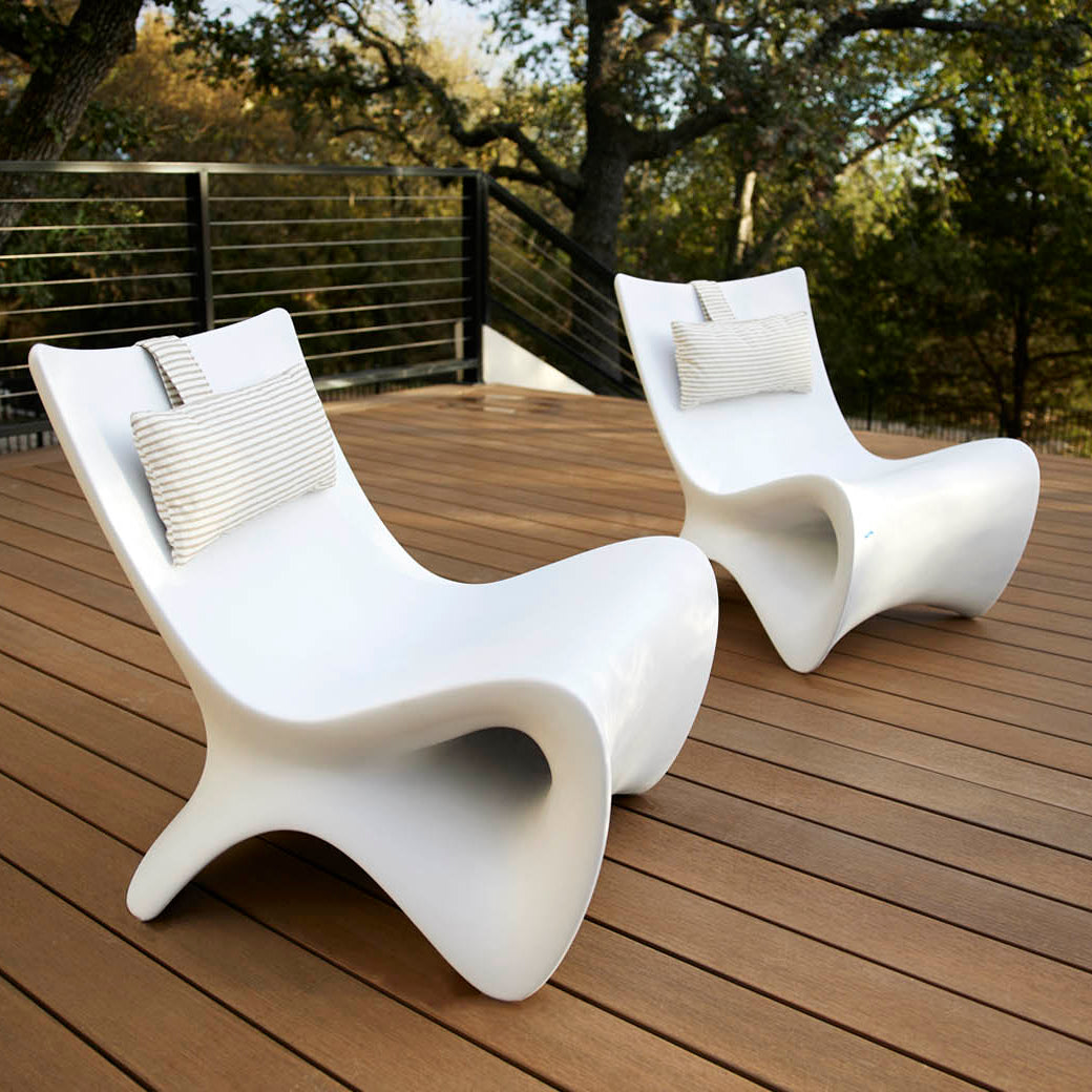 Two Autograph Chairs by Ledge Lounger, in modern white and complete with pillows, enhance a wooden deck surrounded by metal railings, providing breathtaking views of lush green trees for peaceful outdoor relaxation.