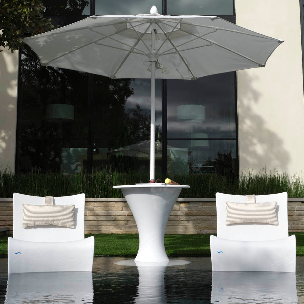 Two Ledge Lounger Autograph Chairs with beige pillows face a sleek white table under a large umbrella outdoors, set by reflective water and enhanced by a contemporary building window in the serene backdrop.