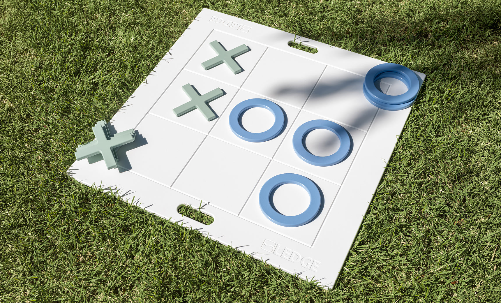 The Ledge Lounger Oversized Patio Games Set features a large white tic-tac-toe board with blue Os and green Xs on green grass. Three Os form a winning line in the center row, making this outdoor game set perfect for backyard fun.