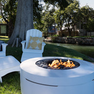 Legacy Adirondack chairs and footrests by Ledge Lounger enhance a grassy area near a peaceful waterway, with a modern white fire pit flickering in the foreground. Trees and houses line the edge, ideal for tranquil patio gatherings.