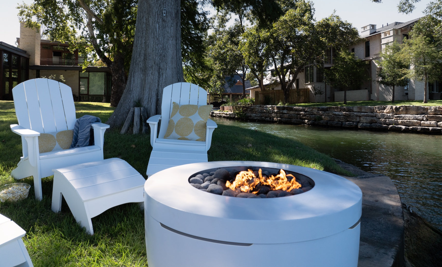 Legacy Adirondack chairs and footrests by Ledge Lounger enhance a grassy area near a peaceful waterway, with a modern white fire pit flickering in the foreground. Trees and houses line the edge, ideal for tranquil patio gatherings.