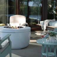 A modern patio setup features a Ledge Lounger Legacy Round Side Table, surrounded by cushioned seating and a fire pit. A candle and glass of rosé grace the table. Large windows reflect greenery and a lake view, creating an inviting outdoor atmosphere.