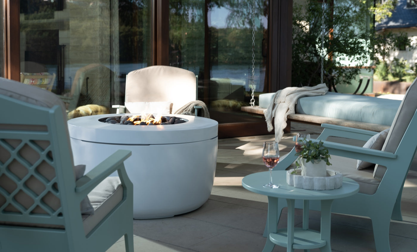 A modern patio setup features a Ledge Lounger Legacy Round Side Table, surrounded by cushioned seating and a fire pit. A candle and glass of rosé grace the table. Large windows reflect greenery and a lake view, creating an inviting outdoor atmosphere.