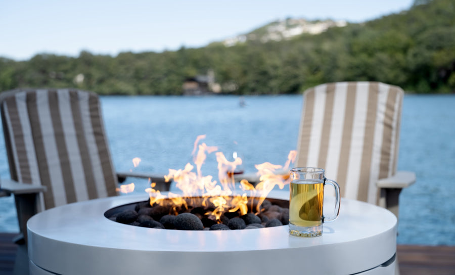 In the foreground, a round fire pit with flames is encircled by smooth stones. A glass mug containing a hot beverage rests on its edge. Two striped deck chairs with Ledge Lounger’s Mainstay Adirondack Cushion for comfort overlook a serene lake and lush green hills.