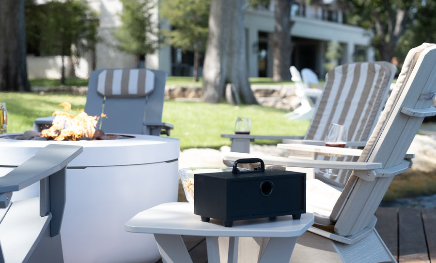 Ledge Lounger’s Mainstay Adirondack Cushions enhance full-body comfort around a modern fire pit. A small table with a black box and drinks sits nearby, set against a serene backdrop of lawn and trees with a house in the distance.