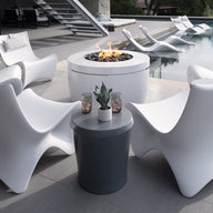 A modern patio features Ledge Loungers sleek white chairs encircling the Halo Aflame Bundle fire pit table. Nearby, two small round tables with plants enhance the setup. Elegant loungers partially submerged in a pool add to the relaxing atmosphere.