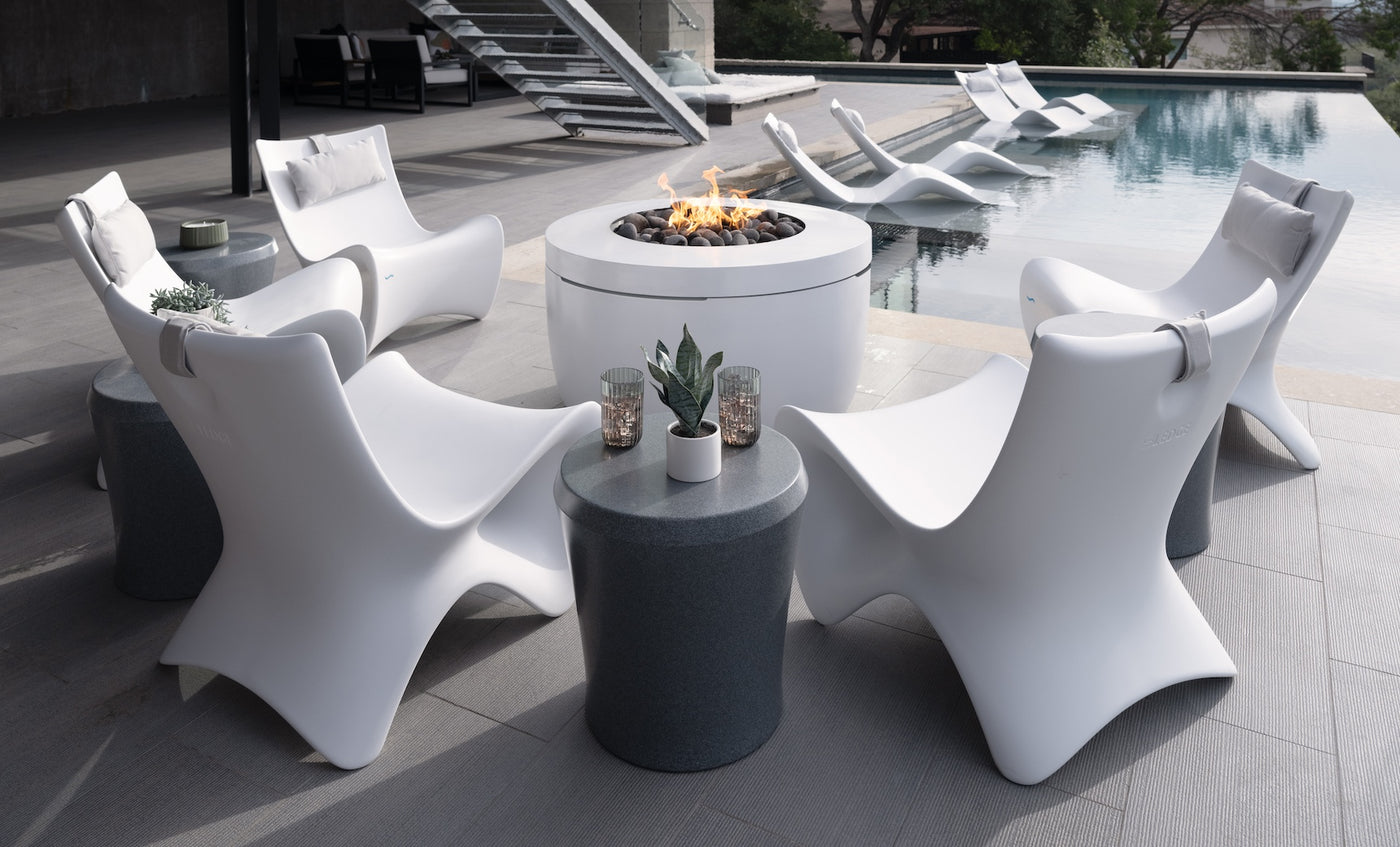 A modern patio features Ledge Loungers sleek white chairs encircling the Halo Aflame Bundle fire pit table. Nearby, two small round tables with plants enhance the setup. Elegant loungers partially submerged in a pool add to the relaxing atmosphere.