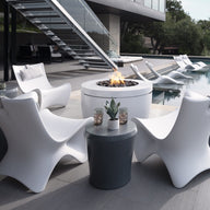 Modern white Autograph Chairs by Ledge Lounger surround a circular fire pit table, creating memorable outdoor moments. A sleek staircase ascends to a rooftop, where an infinity pool with luxury loungers offers relaxation. Nearby, a garden with trees completes this elevated ergonomic oasis.