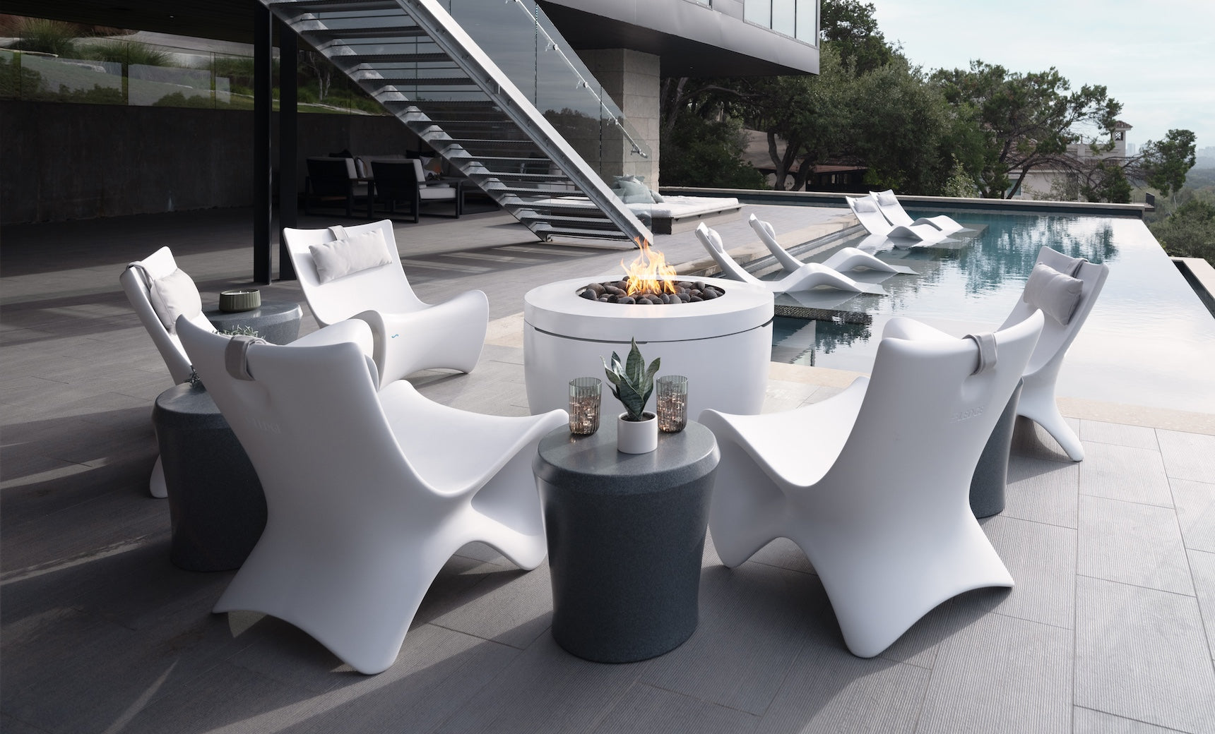 Modern white Autograph Chairs by Ledge Lounger surround a circular fire pit table, creating memorable outdoor moments. A sleek staircase ascends to a rooftop, where an infinity pool with luxury loungers offers relaxation. Nearby, a garden with trees completes this elevated ergonomic oasis.