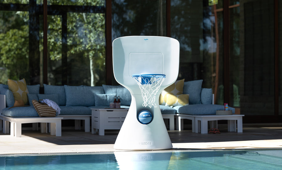 A modern white Ledge Lounger Hoopstr basketball hoop graces the poolside, set against a luxurious outdoor seating area with blue cushions. Sunlight casts shadows, enhancing the peaceful atmosphere, ideal for an impromptu backyard game.