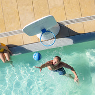 A man shoots a blue basketball toward a poolside hoop from the Ledge Lounger Oversized Patio Games Set, playing in the pool as a woman watches from the edge, viewed from above.