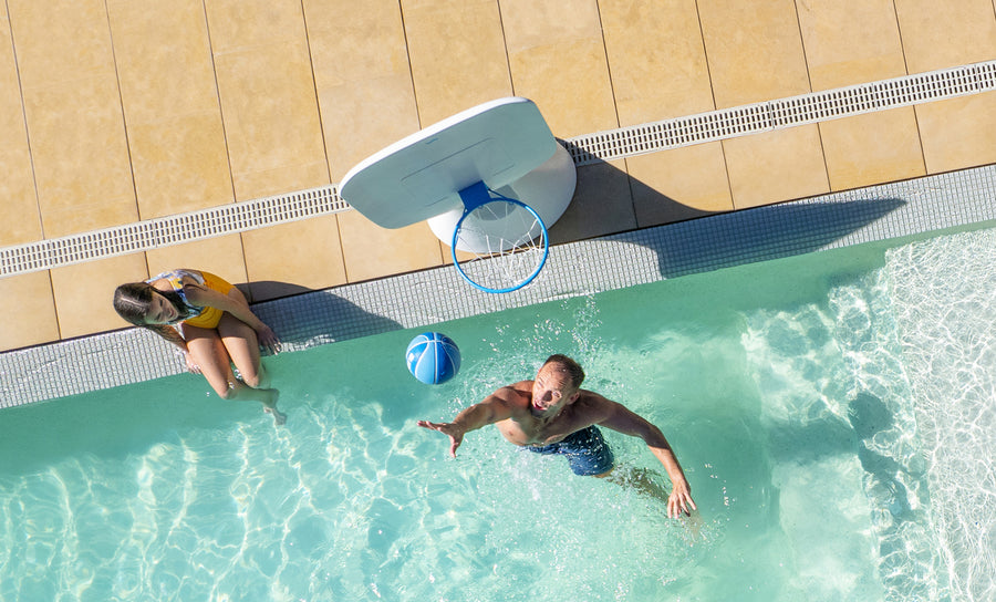 A man shoots a blue basketball toward a poolside hoop from the Ledge Lounger Oversized Patio Games Set, playing in the pool as a woman watches from the edge, viewed from above.