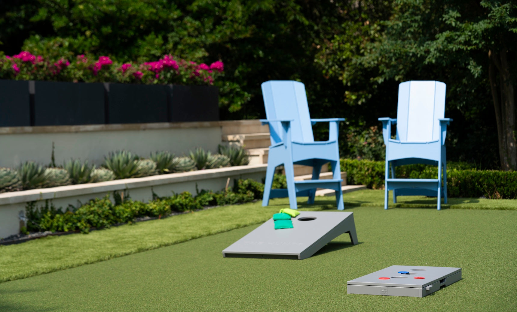 Two light blue Adirondack chairs sit on artificial grass near a Ledge Lounger Cornhole set, with flower beds and green shrubs in the background.