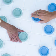 Two hands move circular game pieces on white tiles—one with a light blue piece, the other with teal—part of the Ledge Lounger Oversized Patio Games Set, perfect for all-weather fun or adding to your favorite outdoor game collection.