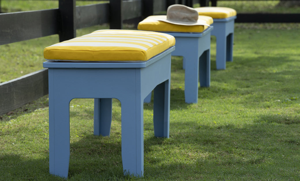 Aligned along a grassy lawn beside a black fence, three light blue Ledge Lounger benches with Mainstay Dining Bench Cushions offer comfort and style. A beige hat rests on the nearest bench, adding casual charm—perfect for those who appreciate customization in outdoor seating.