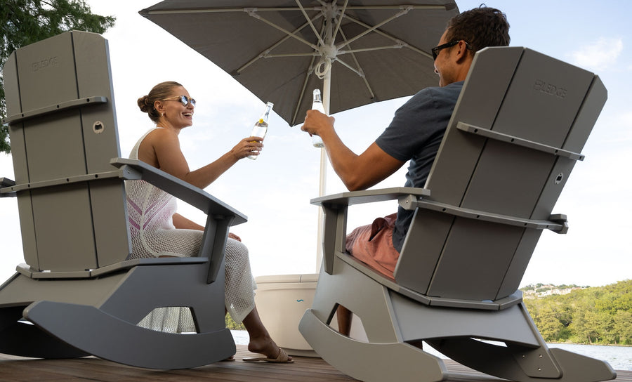 Two people unwind in gray Mainstay Adirondack Rockers by Ledge Lounger on a wooden deck, toasting with bottles under a large umbrella. Theyre near water amid lush greenery, enjoying their sunny and serene outdoor space.