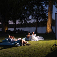 People relaxing on a grassy area by a lake, watching a movie on an outdoor screen at night. They are reclining on Ledge Lounger Laze Pillows and blankets under softly-lit tree canopies. The dimly lit sky enhances the serene ambiance of this outdoor comfort haven.