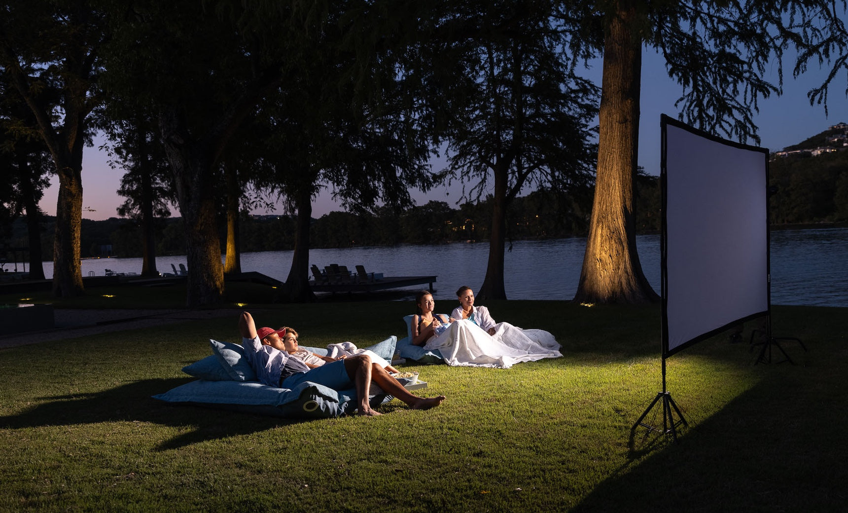 People relaxing on a grassy area by a lake, watching a movie on an outdoor screen at night. They are reclining on Ledge Lounger Laze Pillows and blankets under softly-lit tree canopies. The dimly lit sky enhances the serene ambiance of this outdoor comfort haven.