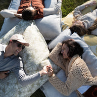 Four people lie on large Laze Pillows by Ledge Lounger in a circle on grass. Two men and two women wear casual, warm clothing, with the pair at the bottom holding hands and smiling, enjoying outdoor comfort.