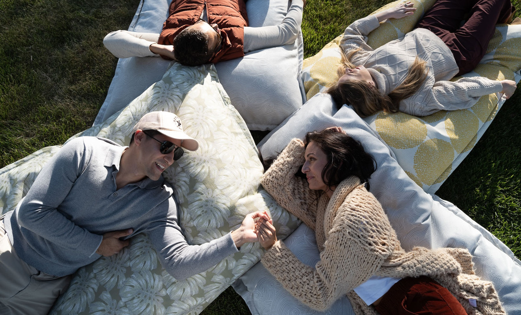 Four people lie on large Laze Pillows by Ledge Lounger in a circle on grass. Two men and two women wear casual, warm clothing, with the pair at the bottom holding hands and smiling, enjoying outdoor comfort.