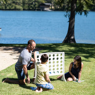 A man and two children enjoy the Ledge Lounger Oversized Patio Games Set outdoors on the grass near a lake, with trees and water in the background on a sunny day.