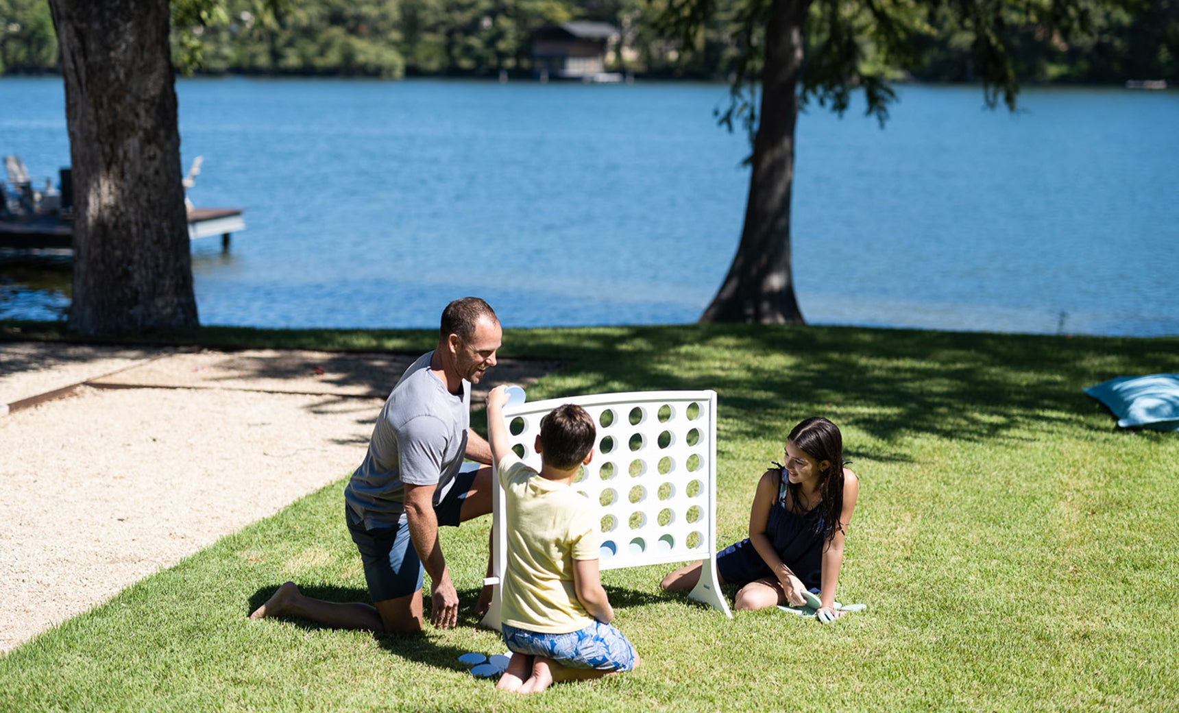 A man and two children enjoy the Ledge Lounger Oversized Patio Games Set outdoors on the grass near a lake, with trees and water in the background on a sunny day.