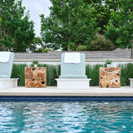 Four Echo Chaise loungers by Ledge Lounger, featuring blue cushions and white towels, are arranged beside a sparkling pool. Wooden side tables are placed between the chairs, set against a backdrop of lush green trees and shrubs for a serene outdoor ambiance.