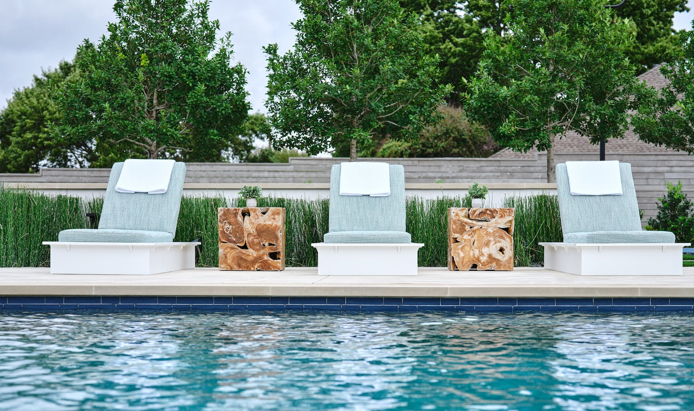 Four Echo Chaise loungers by Ledge Lounger, featuring blue cushions and white towels, are arranged beside a sparkling pool. Wooden side tables are placed between the chairs, set against a backdrop of lush green trees and shrubs for a serene outdoor ambiance.