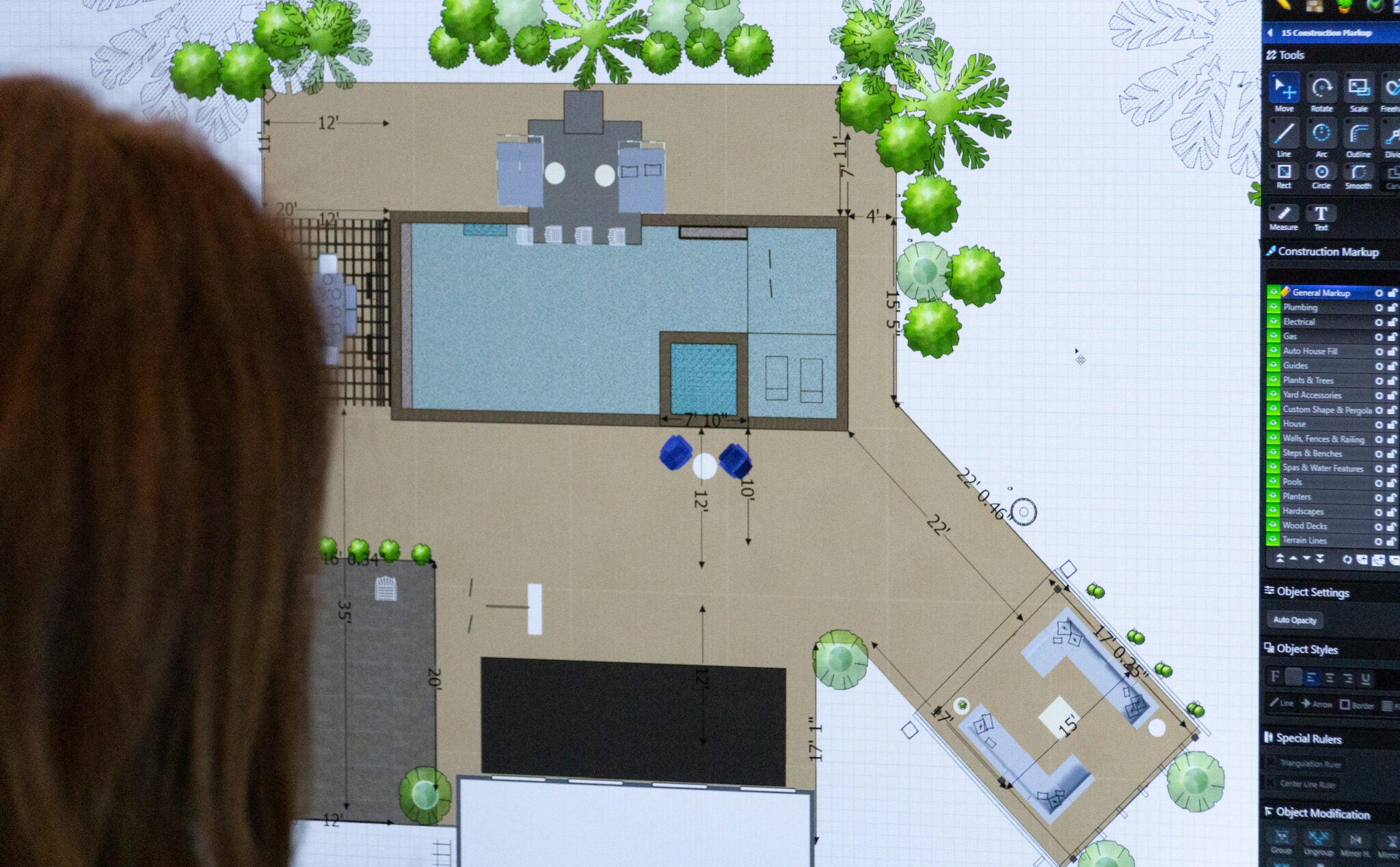 A person looks at an architectural floor plan displayed on a screen. The plan shows a large rectangular pool, surrounding deck area, and outdoor furniture. Green circles represent trees or shrubs, and there are various measurement annotations.