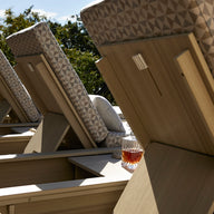 Several Echo Chaise loungers from Ledge Lounger, featuring geometric-patterned cushions, are placed in a serene outdoor space. A glass of brown liquid rests on one lounger’s arm. Tall trees rise in the background under a clear sky.