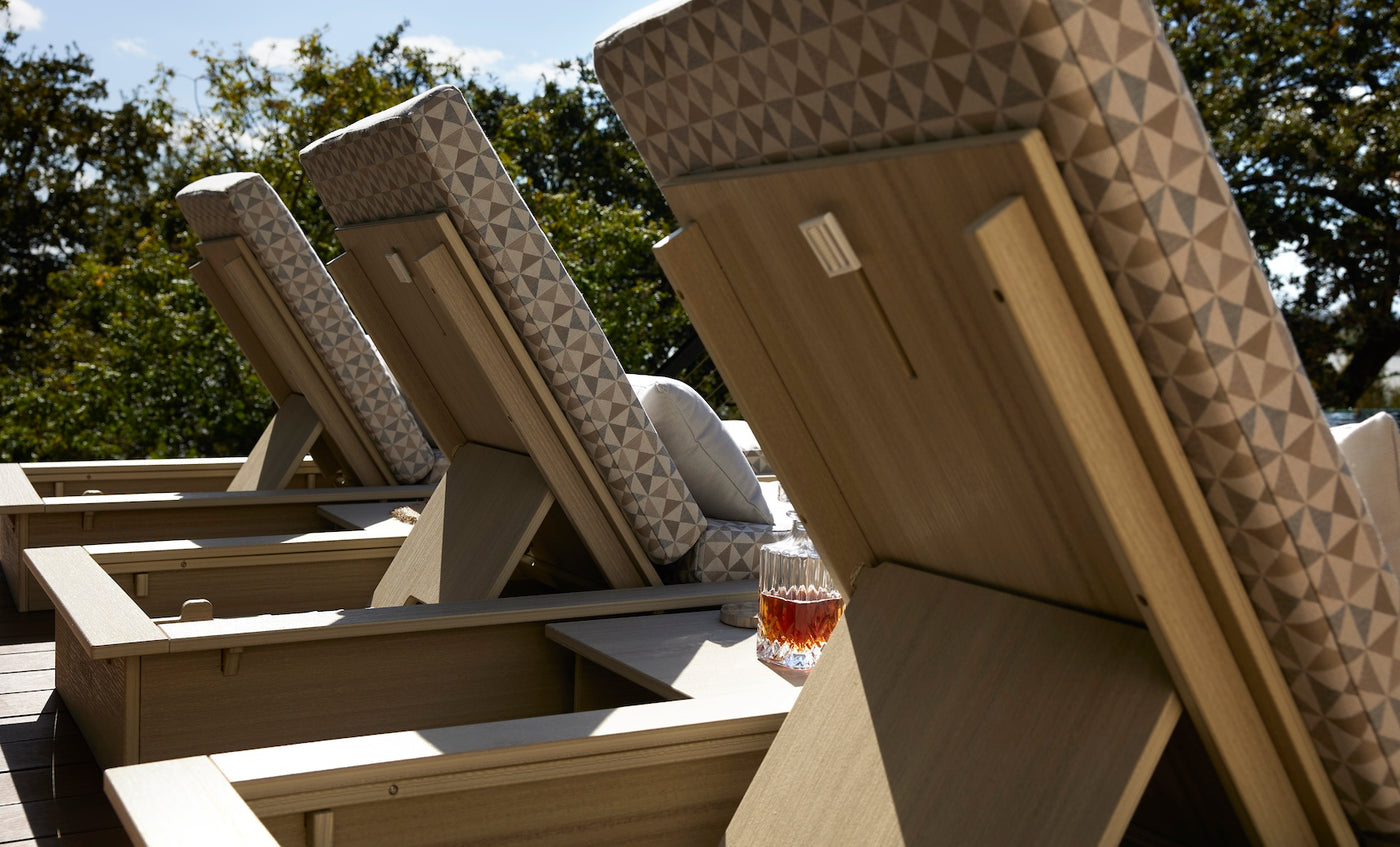 Several Echo Chaise loungers from Ledge Lounger, featuring geometric-patterned cushions, are placed in a serene outdoor space. A glass of brown liquid rests on one lounger’s arm. Tall trees rise in the background under a clear sky.