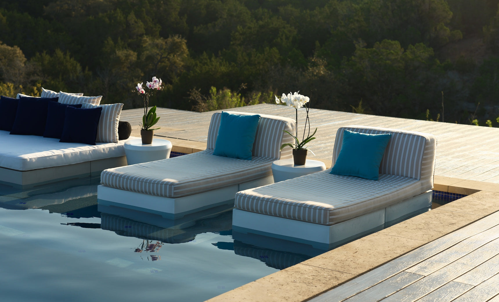 Two Ledge Lounger Affinity 2 Piece Sun Chairs with blue outdoor cushions and small side tables topped with potted orchids sit by a pool, surrounded by trees and bathed in gentle sunlight.